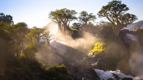 Sonnenaufgang über einem Wasserfall mit Baobab Bäumen im Vordergrund.