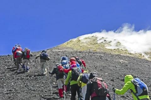 Eine Gruppe Wanderer besteigt einen Vulkan. Der Boden ist steinig. Aus dem Vulkan kommt Rauch.