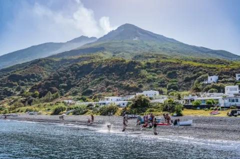 Menschen baden am Strand. Im Hintergrund sind ein paar Häuser und weit hinten ein Vulkan.