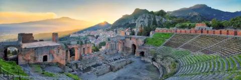 Ein Theater mit einer rund gebauten Tribüne. Dahinter ist ein Dorf und Berge. Die Sonne geht unter.