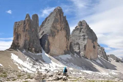 Wanderer mit Rucksack steht auf alpinem Gelände vor drei hohen und massiven Felsen bei leicht bewölktem Himmel