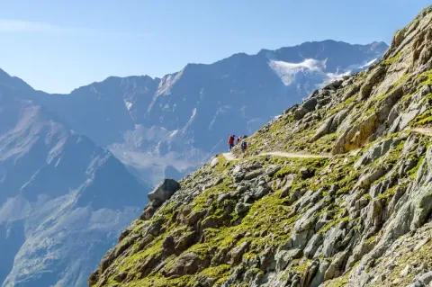 Zwei Personen laufen auf dem Wanderweg entlang eines steilen Berghangs durch Berglandschaft