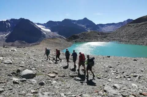 Gruppe von sechs Personen läuft durch Gerölllandschaft vorbei an einem blauen Bergsee