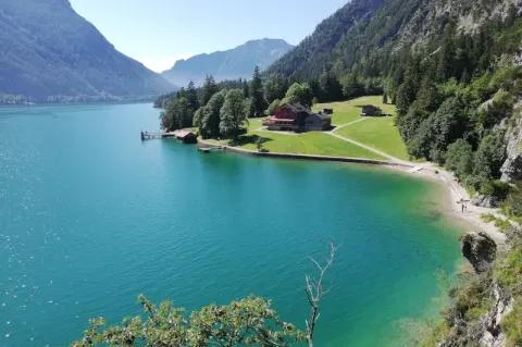 Panorama eines blauen Sees mit Uferpromenade, Wiesen und bewaldeten Berghängen
