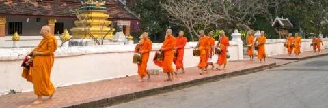 Mönchen in orangener Kleidung laufen auf Bürgersteig am Straßenrand hintereinander entlang