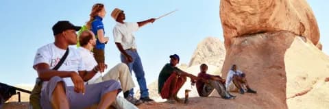 Ein Reiseleiter mit weißem T-Shirt und Hut zeigt mit einem Stock auf eine felsige Formation. Eine Frau steht neben ihm und mehrere Männer sitzen am Boden um ihn herum.