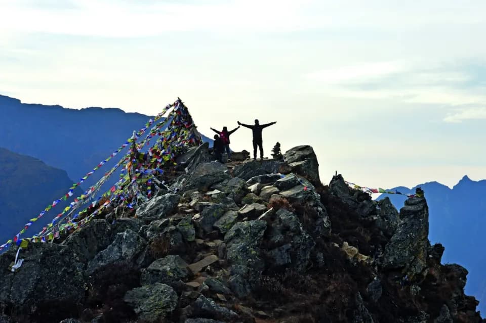 Drei Wanderer stehen auf einem Gipfel und heben die Arme. Es wehen Gebetsfahnen um den Gipfel.