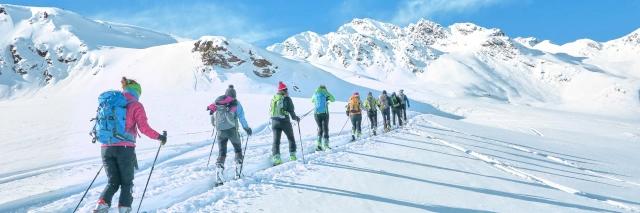 Neun Personen mit Skiern laufen durch eine schneebedeckte Berglandschaft unter blaue Himmel