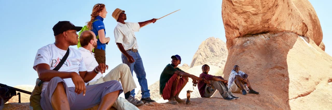 Ein Reiseleiter mit weißem T-Shirt und Hut zeigt mit einem Stock auf eine felsige Formation. Eine Frau steht neben ihm und mehrere Männer sitzen am Boden um ihn herum.