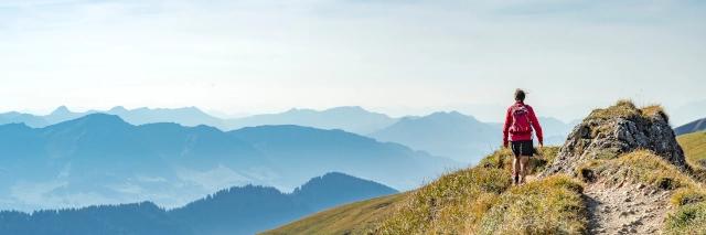 Frau mit Rucksack läuft auf einem leichten Grat mit Wiesen mit Bergen im Hintergrund