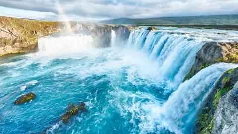 Panoramablick auf einen mehrstufigen Wasserfall mit türkisfarbenem Wasser und einem Regenbogen.