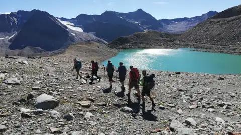 Wandergruppe auf einem Bergpfad neben einem türkisfarbenen See mit Bergen und Gletscher im Hintergrund.